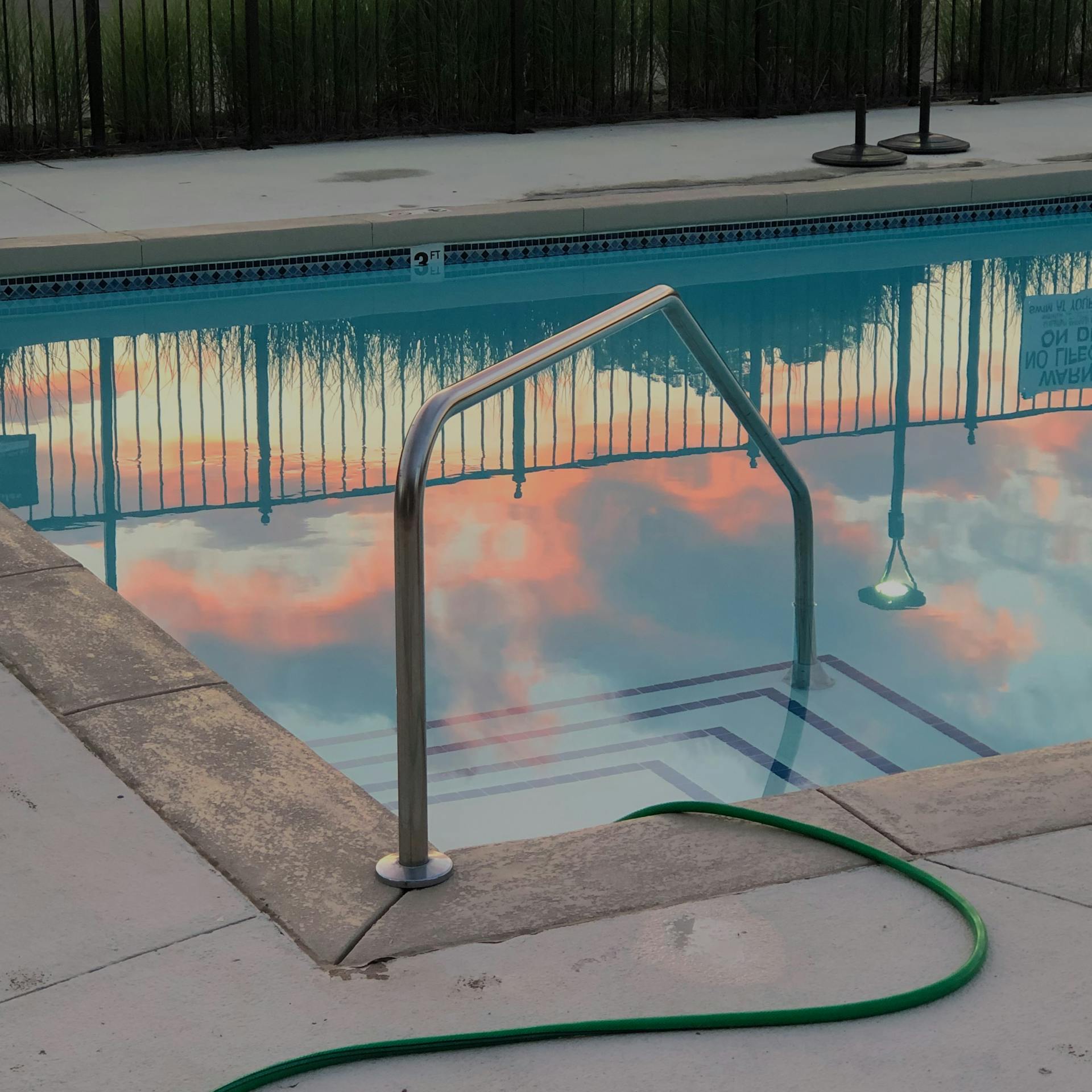 Empty small swimming pool with metal handrail and hose in backyard next to fence in evening